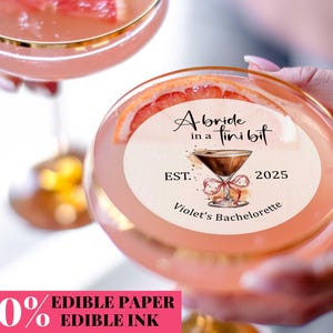 May include: A pink cocktail with a gold rim in a glass. A pink and white round sticker with the text "A bride in a tiny bit EST. 2025 Violet's Bachelorette" is on top of the glass.