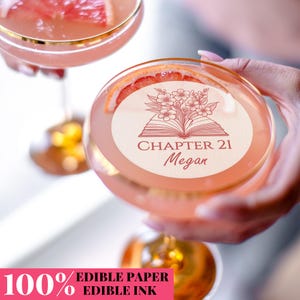 May include: Two pink cocktails in stemmed glasses, garnished with grapefruit slices. One glass is held up, revealing a circular edible paper with a floral design, an open book, and the text "CHAPTER 21 Megan". The image also includes the text "100% EDIBLE PAPER EDIBLE INK".