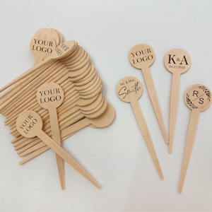 Puede incluir: Palitos de madera para remover con tapas circulares. Algunos están grabados con "YOUR LOGO", otros con "K&A 06.17.2026", "Mr. & Mrs. Smith" y un monograma con "R" y "S" dentro de una corona. Los palitos son de color marrón claro.