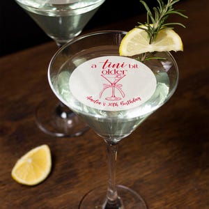 Puede incluir: Una pegatina redonda blanca con texto rojo que dice "a  tini bit older" y una ilustraci&oacute;n roja de una copa de martini. La pegatina est&aacute; colocada en una copa de martini. El texto "Andie's 30th Birthday" tambi&eacute;n est&aacute; en la pegatina.