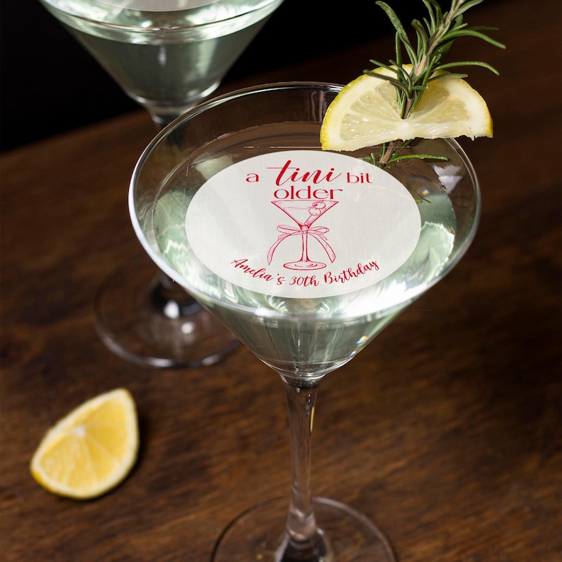 Puede incluir: Una pegatina redonda blanca con texto rojo que dice "a  tini bit older" y una ilustraci&oacute;n roja de una copa de martini. La pegatina est&aacute; colocada en una copa de martini. El texto "Andie's 30th Birthday" tambi&eacute;n est&aacute; en la pegatina.