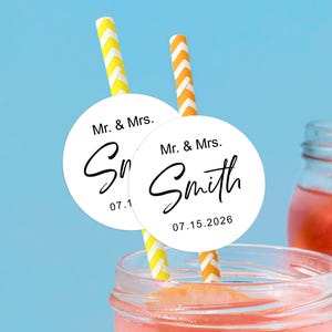 Puede incluir: Dos etiquetas circulares blancas para bebidas con el texto «Sr. y Sra. Smith» y la fecha «07.15.2026» están adheridas a pajitas a rayas amarillas y naranjas. Las pajitas están en un vaso de líquido rosa, con un fondo azul.