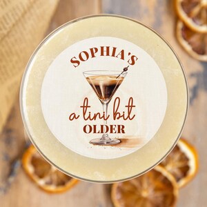Puede incluir: Una pegatina redonda blanca con texto marr&oacute;n que dice "Sophia's a  tini bit older" y un dibujo de una copa de c&oacute;ctel con un l&iacute;quido marr&oacute;n. La pegatina est&aacute; encima de un vaso de l&iacute;quido amarillo claro.