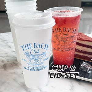May include: A clear plastic cup and lid set with the text "THE BACH CLUB" in blue. A tall glass with a red beverage and a slice of red velvet cake are also visible. The words "CUP & LID SET" are printed on the image.