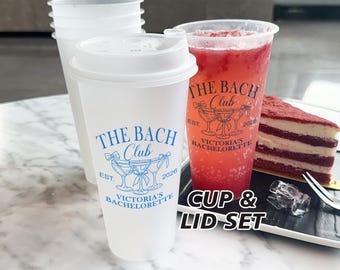 Puede incluir: Un juego de vasos y tapas de plástico transparente, cada uno con la impresión "THE BACH CLUB" en azul. También se ve un vaso alto lleno de una bebida roja y una porción de pastel de terciopelo rojo. El texto "CUP & LID SET" está impreso.