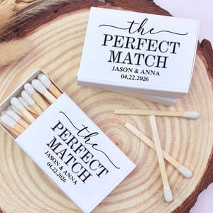 May include: White matchboxes with the text "the PERFECT MATCH" and names and a date. The matchboxes are open, revealing the matchsticks with white tips. Three loose matches are also visible. The items are on a wooden surface.