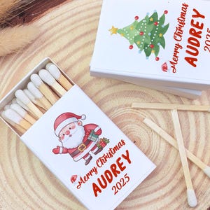 May include: White matchboxes with Christmas-themed illustrations. One box features Santa Claus, the other a Christmas tree. Both boxes have the text "Merry Christmas AUDREY 2025" in red. Matches with white tips are visible.