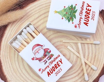 May include: White matchboxes with Christmas-themed illustrations. One box features Santa Claus, the other a Christmas tree. Both boxes have the text "Merry Christmas AUDREY 2025" in red. Matches with white tips are visible.