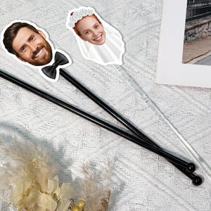 Puede incluir: Dos agitadores de cóctel. Uno negro con la foto de un hombre sonriente con barba y pajarita negra. El otro es transparente con la foto de una mujer con velo. Los agitadores están sobre una superficie blanca texturizada.