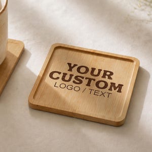 K&ouml;nnte beinhalten: Ein quadratischer Bambusuntersetzer mit abgerundeten Ecken und einem erh&ouml;hten Rand. In der Mitte des Untersetzers ist der Text "YOUR CUSTOM LOGO / TEXT" in einer dunkelbraunen Schrift eingraviert. Die Holzmaserung ist auf der Oberfl&auml;che sichtbar.