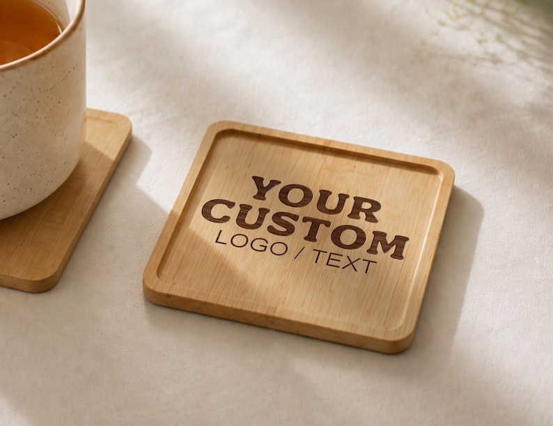 K&ouml;nnte beinhalten: Ein quadratischer Bambusuntersetzer mit abgerundeten Ecken und einem erh&ouml;hten Rand. In der Mitte des Untersetzers ist der Text "YOUR CUSTOM LOGO / TEXT" in einer dunkelbraunen Schrift eingraviert. Die Holzmaserung ist auf der Oberfl&auml;che sichtbar.