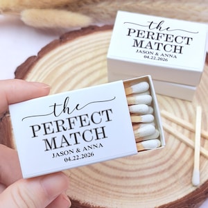 May include: White matchboxes with the text "the PERFECT MATCH" and the names "JASON & ANNA" and the date "04.22.2026" printed in black. The matchbox is open, revealing matches with white tips. The image is on a wooden surface.