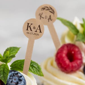 May include: Two wooden cake toppers with the initials "K & A" and the date "06.17.2026" are displayed. The toppers are placed in a dessert with red raspberries, blueberries, and mint leaves. The background is blurred, highlighting the toppers.