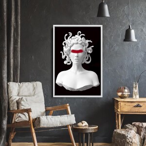 May include: A framed art print depicting a white bust of Medusa with a red band across the eyes, set against a black backdrop. The artwork is displayed in a room with a wooden chair, side table, and dark walls.