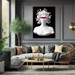 May include: A white bust of Medusa with a red stripe across the eyes, set against a black background. The artwork is displayed in a modern living room with a grey sofa, gold cushions, and a wooden coffee table.