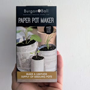 May include: A black and gold box for a paper pot maker. The box shows seedlings in newspaper pots. Text on the box reads "PAPER POT MAKER" and "MAKE A LIMITLESS SUPPLY OF SEEDLING POTS." The brand name is "Burgon & Ball."