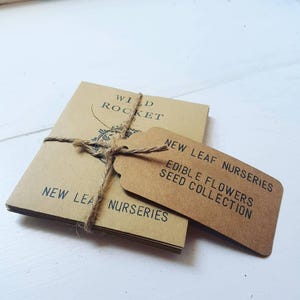 May include: A stack of brown paper seed packets tied with twine and a brown paper tag that reads "New Leaf Nurseries Edible Flowers Seed Collection". The top packet has the text "Wild Rocket" printed on it.
