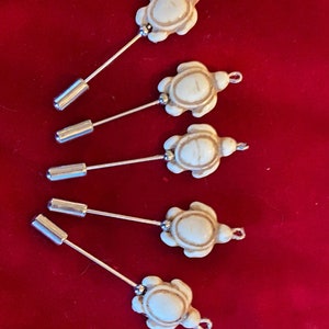 Puede incluir: Cinco alfileres blancos en forma de tortuga con tallos de metal plateado. Las tortugas tienen un patrón de mármol y están unidas a los tallos con un lazo de metal plateado.