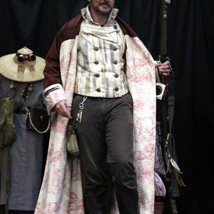May include: A man wearing a long, white and pink patterned coat with a burgundy trim. He is also wearing a striped vest, black pants, and black boots. He is holding a long, wooden staff.