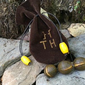 May include: A brown velvet pouch with the letters "HTH" embroidered on it. The pouch is tied with a black cord and has yellow wooden beads attached to it. There are three brown seed pods on the ground next to the pouch.
