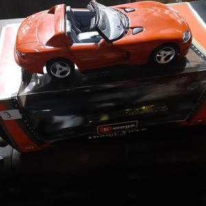 Puede incluir: Un coche descapotable de juguete rojo con ruedas plateadas e interior negro. El coche es un modelo de Dodge Viper RT/10. El coche está encima de una caja negra con el logo de Burago.