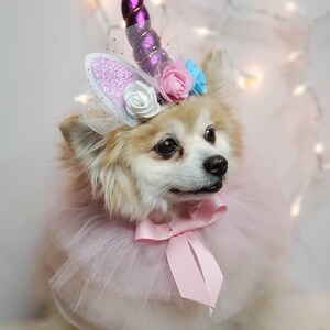 May include: A small, white dog wearing a pink tulle tutu and a unicorn headband with purple, pink, and blue flowers. The headband has a sparkly white ear and a purple horn.