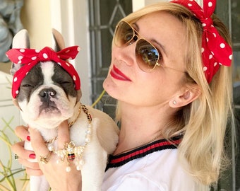 Könnte beinhalten: Eine Frau mit einem roten und weißen gepunkteten Stirnband und Sonnenbrille hält einen weißen französischen Bulldoggen-Welpen, der ein passendes rotes und weißes gepunktetes Stirnband trägt. Der Welpe trägt eine goldene Kette mit einem Perlen- und Bienenanhänger.