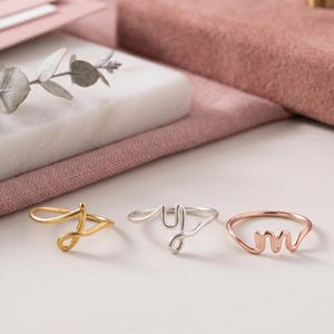 Op de afbeelding: Drie dunne metalen ringen, elk voorzien van een enkele cursieve letter gevormd uit draad. De eerste ring is goudkleurig met de letter J, de tweede is zilverkleurig met de letter y en de derde is roségoudkleurig met de letter m.