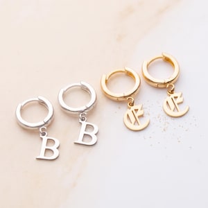 May include: Two pairs of hoop earrings with dangling letter charms. One pair is silver-toned with the letter B, and the other pair is gold-toned with a stylised letter E. Each earring features a circular hoop with a hinged closure and a small charm.