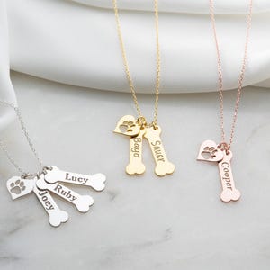 May include: Three necklaces in silver, gold, and rose gold tones. Each features a delicate chain with dangling charms: a heart with a cutout paw print and one or more bone-shaped pendants engraved with the names Joey, Lucy, Ruby, Bayo, Sauer, or Cooper.