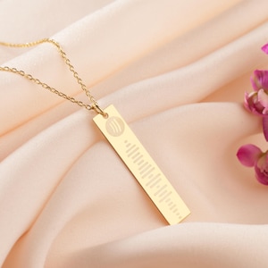 Op de afbeelding: Een goudkleurige hangerketting met een rechthoekige hanger. De hanger heeft een muziek golfvorm ontwerp en een cirkelvormig logo. De ketting wordt getoond op een zachte, perzik kleurige stof.