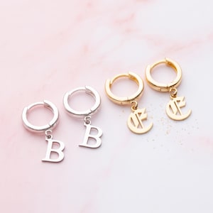 May include: Two pairs of hoop earrings, one silver-toned and one gold-toned. The silver pair features a dangling letter B charm, while the gold pair features a dangling letter charm with a stylised, gothic-inspired font design.