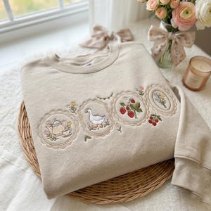 Könnte beinhalten: Beiges Sweatshirt mit Rundhalsausschnitt und gestickten Blumen- und Naturmotiven. Die Stickerei umfasst eine Teekanne, eine Gans, Erdbeeren und Blumen in dekorativen Rahmen. Das Sweatshirt liegt auf einem geflochtenen Tablett.