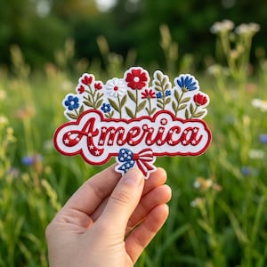 Op de afbeelding: Een geborduurde patch met het woord America in rode schrijfletters met witte sterren, bekroond met diverse rode, witte en blauwe bloemen en groene stelen. Een kleine blauwe strik met witte sterren is in het midden onder de tekst bevestigd.