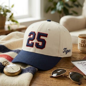 Op de afbeelding: Een crèmekleurige baseballcap met een marineblauwe klep en het nummer 25 in marineblauw met rode rand. Het woord "Tiger" is op de zijkant geborduurd. Een vintage zakhorloge, zonnebril en een mok zijn ook zichtbaar.