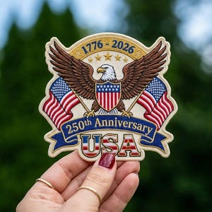 May include: A commemorative embroidered patch featuring a bald eagle with spread wings, centered above a shield with red and white stripes. Two American flags flank the eagle. Text reads 1776-2026, 250th Anniversary, and USA in bold letters.