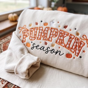 May include: A cream-coloured sweatshirt featuring embroidered text that reads PUMPKIN season. The word PUMPKIN is in orange and white gingham block letters, while season is in black script. The design includes small pumpkins, leaves, stars, and a ghost.