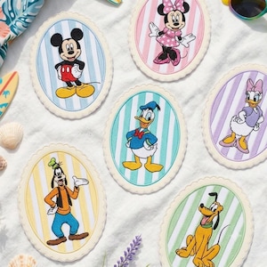 Op de afbeelding: Vijf ovale stoffen onderzetters met geborduurde Disney-figuren: Mickey Mouse op blauwe strepen, Minnie Mouse op roze strepen, Donald Duck op turquoise strepen, Daisy Duck op paarse strepen, Goofy op gele strepen en Pluto op groene strepen.