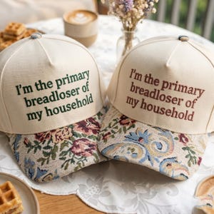 May include: Two cream-colored baseball caps with floral patterned brims. The caps have the embroidered text, "I'm the primary breadloser of my household" in green and brown. The caps are on a table with food and flowers.