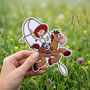 May include: An embroidered patch featuring a cartoon cowgirl with red hair and a white shirt riding a brown horse. The cowgirl is holding a lasso above her head. The patch has a white border and depicts the characters in vibrant colors.