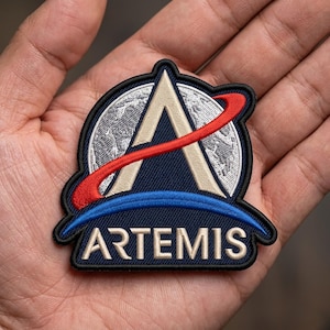 May include: Embroidered patch with the Artemis program logo. The patch has a navy blue background with a white and beige stylized 'A' shape, a red arc, and a blue arc. The word "ARTEMIS" is embroidered in white.