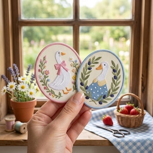 May include: Two oval embroidered patches featuring white geese. The left patch has a goose with a pink bow surrounded by colorful flowers and a pink border. The right patch shows a goose in a blue polka dot dress with olive branches and a blue border.