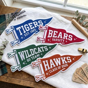 May include: Four felt pennants in blue, red, green, and orange, each featuring white embroidered text. The blue pennant reads TIGERS EST. 1985, the red reads BEARS VARSITY, the green reads WILDCATS GO TEAM!, and the orange reads HAWKS CLASS OF 2026.