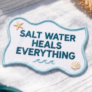 May include: A white embroidered patch with a light blue scalloped border. The text reads SALT WATER HEALS EVERYTHING in dark teal thread. A small beige starfish is in the top left corner, and a beige seashell is in the bottom right corner.