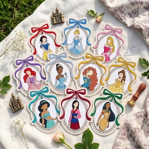 May include: A collection of ten oval-shaped embroidered patches, each featuring a different female character in a white frame topped with a coloured ribbon bow. The characters include Snow White, Cinderella, Aurora, Ariel, Tiana, Merida, Belle, Jasmine, Mulan, and Pocahontas, all rendered in a stylised, faceless illustration format.