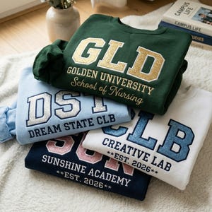 May include: A stack of sweatshirts in various colors: green, light blue, white, and navy. Each sweatshirt features collegiate-style lettering and text, including "GLD", "DST", "CLB", and "Sunshine Academy". The sweatshirts are on a white, textured surface.
