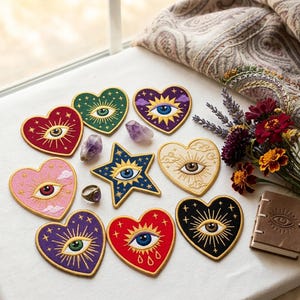 May include: A collection of embroidered patches in heart and star shapes, each featuring a central eye motif with radiating lines, clouds, and stars. The patches are in various colors including red, green, purple, pink, black, and cream.
