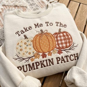 May include: A cream-colored crewneck sweatshirt featuring brown embroidered text that reads Take Me To The PUMPKIN PATCH. The design includes three pumpkins with different patterns: floral, polka dot, and gingham, accented by embroidered autumn leaves.