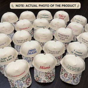 May include: A collection of cream-colored baseball caps with various embroidered phrases in different colors. The caps have floral patterned brims. Phrases include "I'm fine it's fine", "Someone's mean ass wife", and "Mimi".