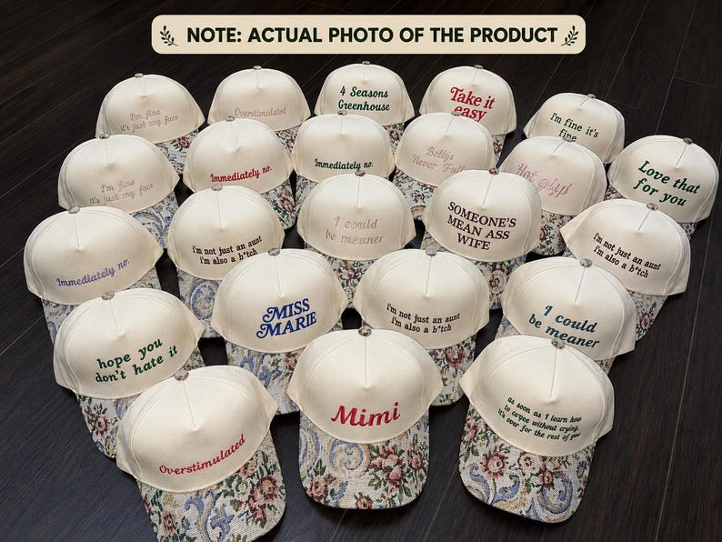 May include: A collection of cream-colored baseball caps with various embroidered phrases in different colors. The caps have floral patterned brims. Phrases include "I'm fine it's fine", "Someone's mean ass wife", and "Mimi".
